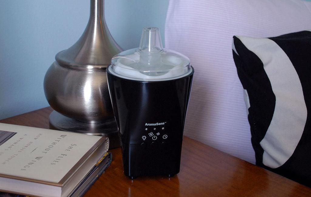 Black AromaSens ultrasonic diffuser on nightstand with books and lamp base