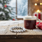 A steaming mug of hot chocolate with marshmallows on a wooden table, with a Wyndmere aromatherapy bracelet and a Christmas tree and lights in the background.