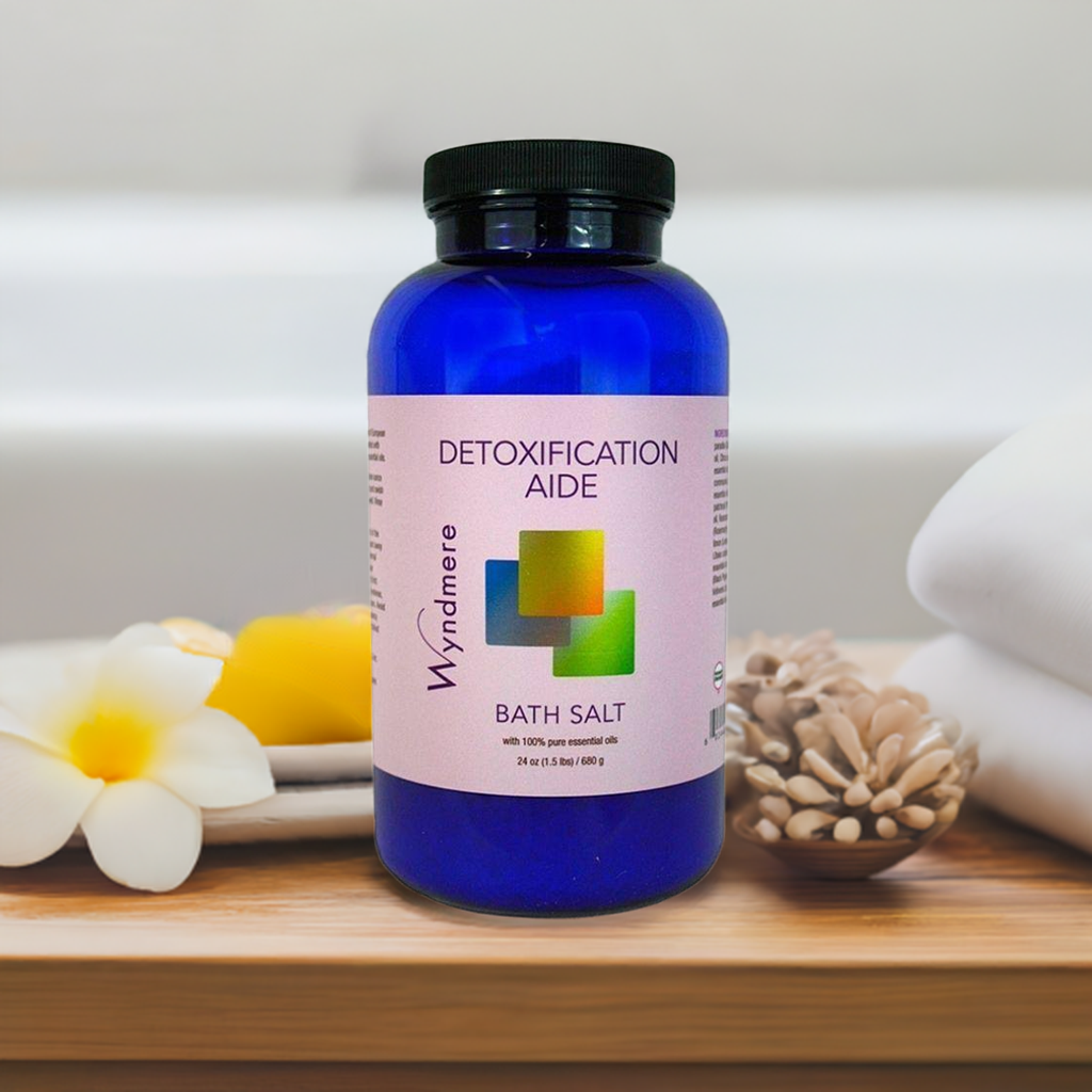 Blue bottle of Wyndmere Detoxification Aide Bath Salt on a wooden surface with flowers and towels in the background.
