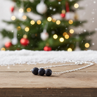 Wyndmere necklace with three dark beads on a wooden surface in front of a Christmas tree with lights.