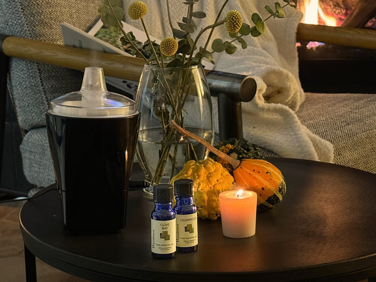 Wyndmere Clove Bud and Cedarwood bottles beside a diffuser, with a candle, gourds and a cozy throw by the fire