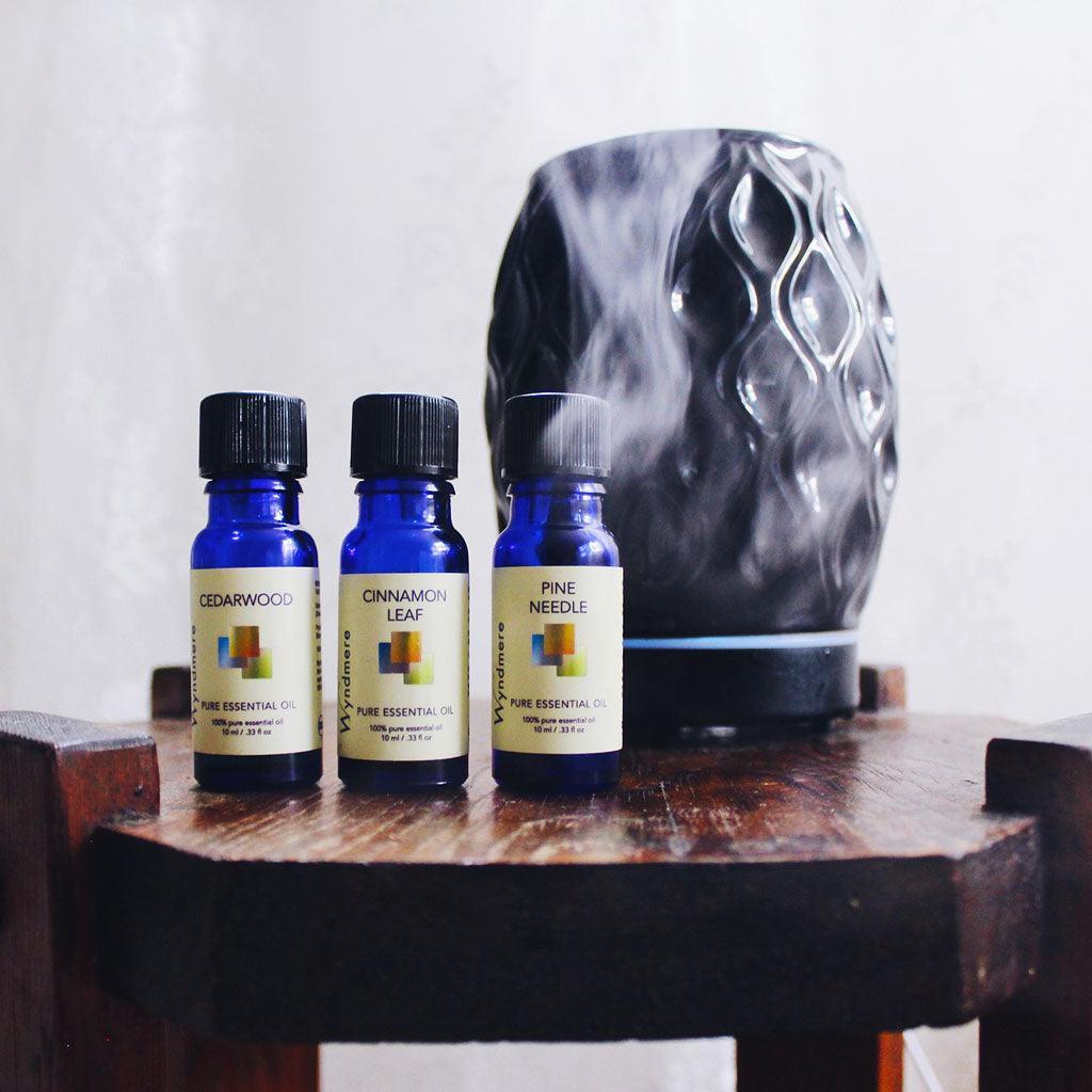 Black AromaVase ultrasonic diffuser on wood table showing vapors flowing over bottles of Wyndmere Cedarwood, Cinnamon, and Pine essential oils.