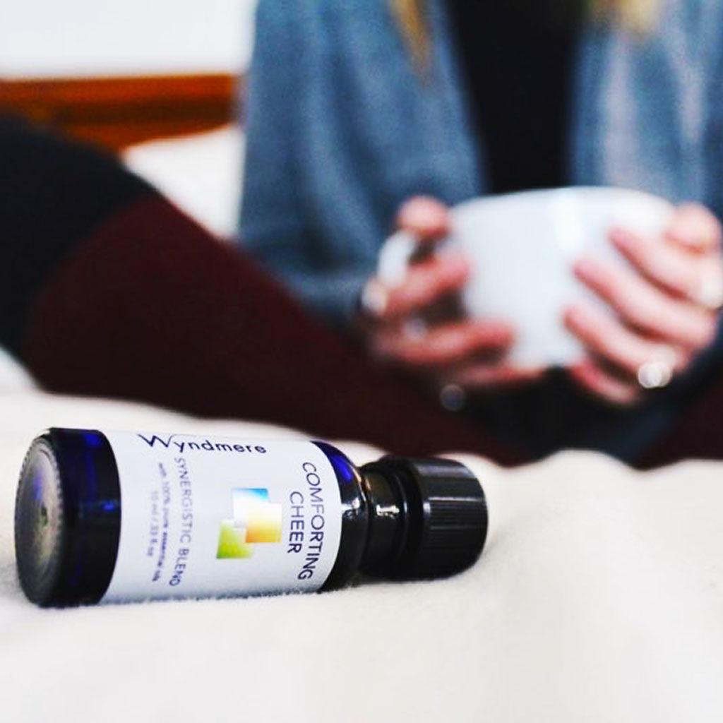 Woman sitting on bed with a warm cup of coffee and a bottle of Wyndmere Comforting Cheer